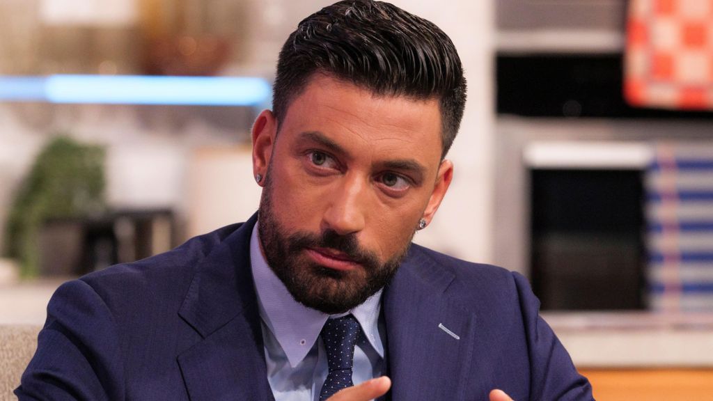 Giovanni Pernice on ITV's Lorraine show, he looks serious and is wearing a navy blue suit, a navy tie with white dots and a light blue shirt.