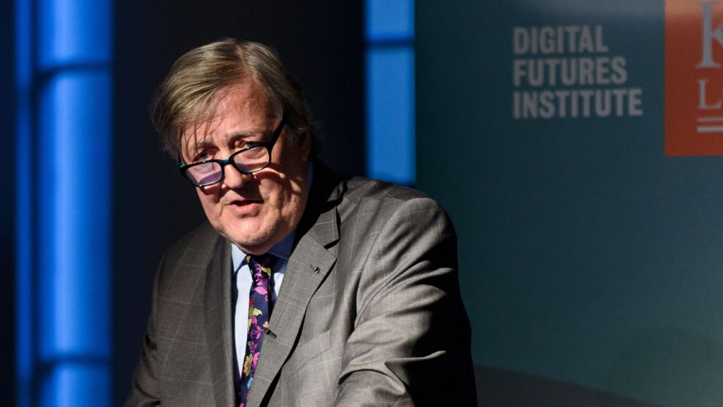 Stephen Fry, standing at a lectern in a suit and tie