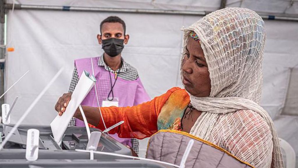 A woman in a white headscarf and orange top puts a ballot paper in ballot box. An official wearing a lilac tabard and facemask watches on.