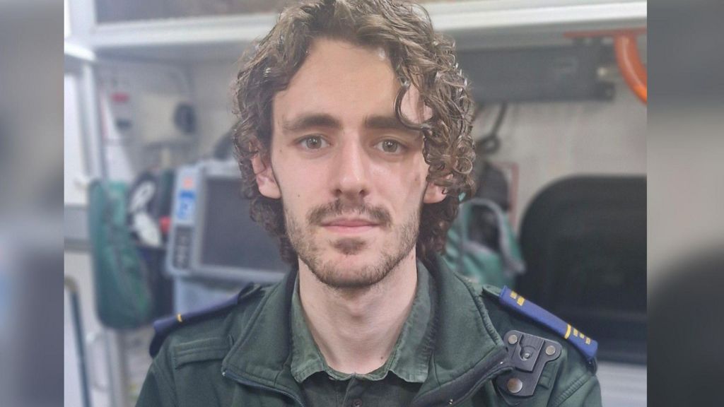Hervey Jenkins, a young man with curly brown hair, looks at the camera with a straight face. He wears a green medical uniform.