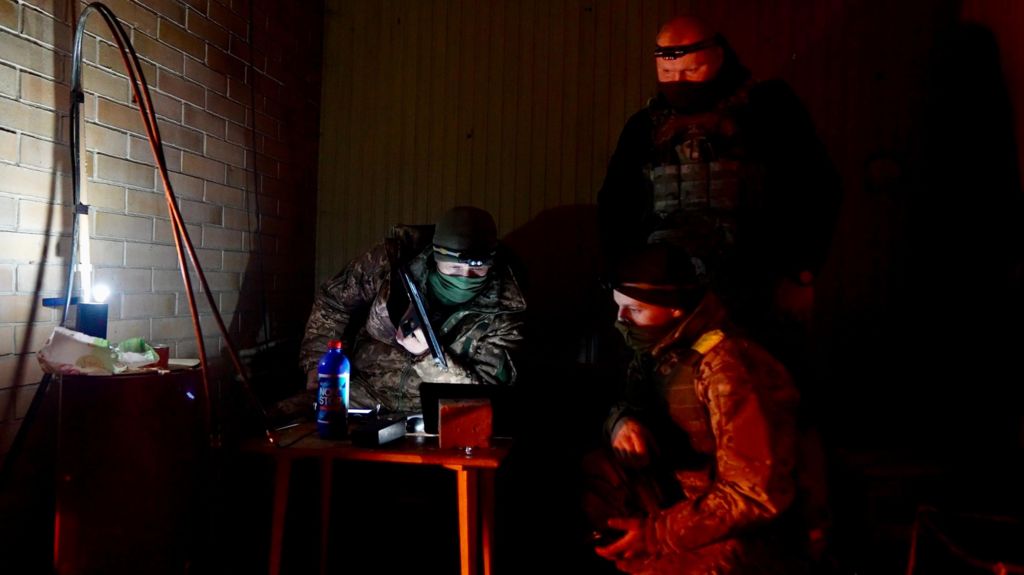 Two masked men look at a computer screen with another masked man standing over him, in a dark room lit up by one lamp