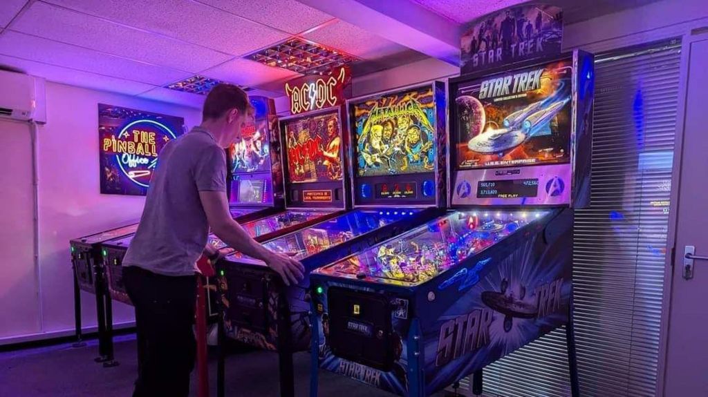 A man in a grey T-shirt has both hands on the sides of a pinball machine, one in a line of four