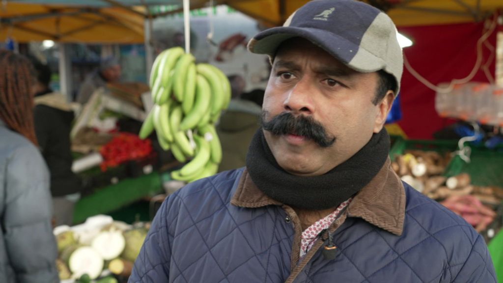 Jose Joseph, wearing a baseball cap, stands in front of his market stall, a large bunch of bananas hanging in the background