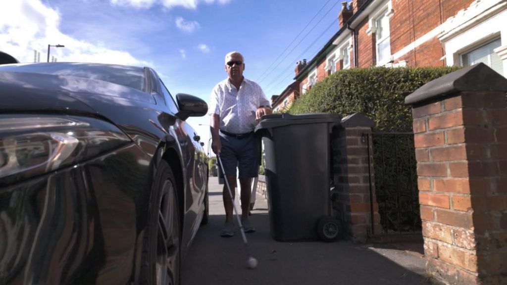 A man with a white cane trying to squeeze past a car parked on a pavement and a wheelie bin. 