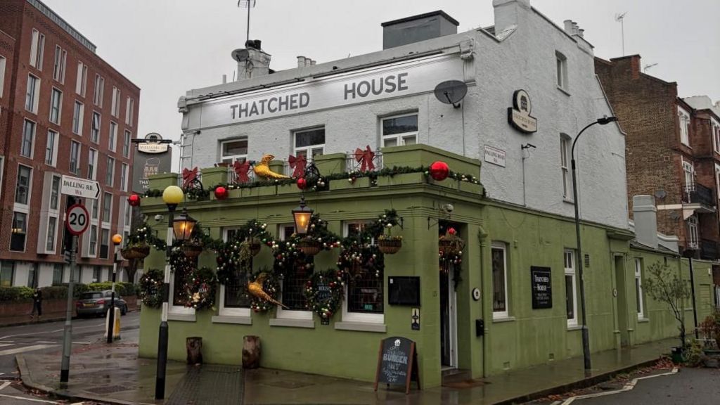 A pub with a green brick first floor and white top floor. Signage reads "Thatched House and the windows are decorated with festive garlands and baubles. 