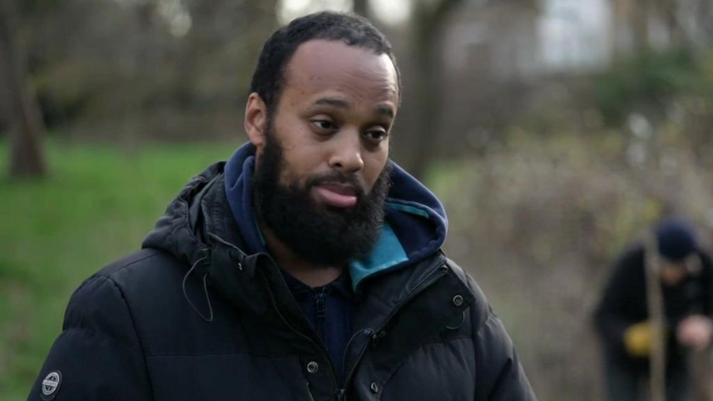 Head-and-shoulders image of a Mahamed Hashi, seen in a dark jacket in a park, with another person planting a young tree in the background.