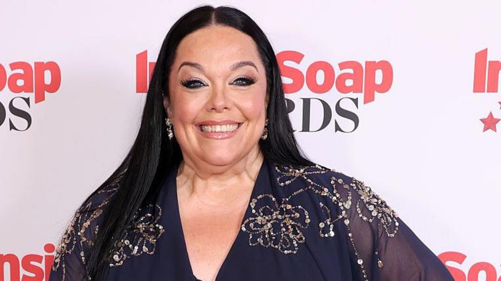 A photo of Lisa Riley from the shoulders up. She has long dark hair and is wearing a blue dress with beaded embellishments, and is standing in front of a background with the logo for the Soap Awards. She is smiling at the camera.