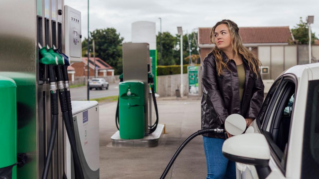 A woman wearing blue jeans and a brown coat fills up her white hatchback car at a petrol station.