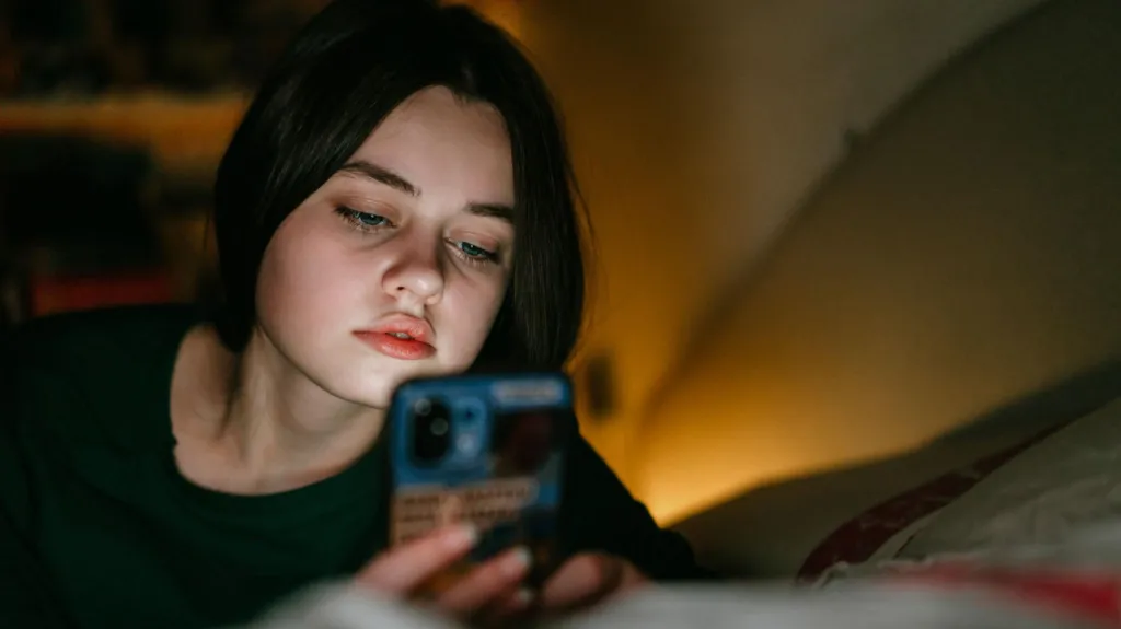 A woman looks at her phone lying on the bed in a low light.