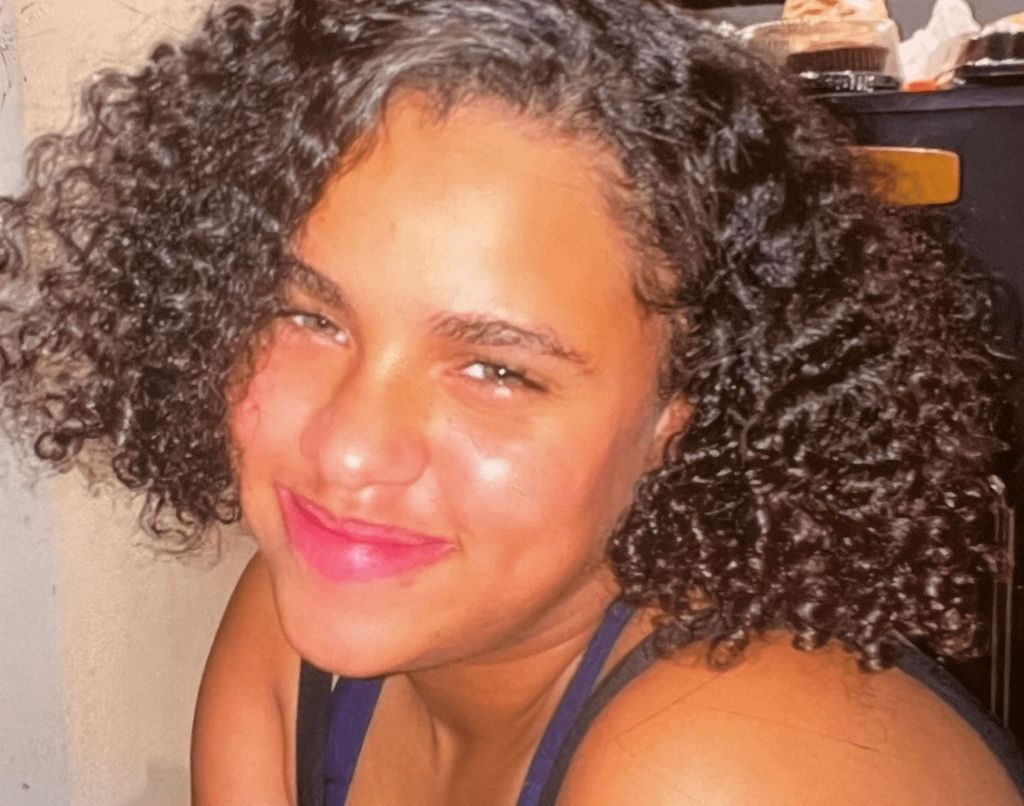 Emily, smiling at the camera. She has brown curly hair and wears a vest top. 