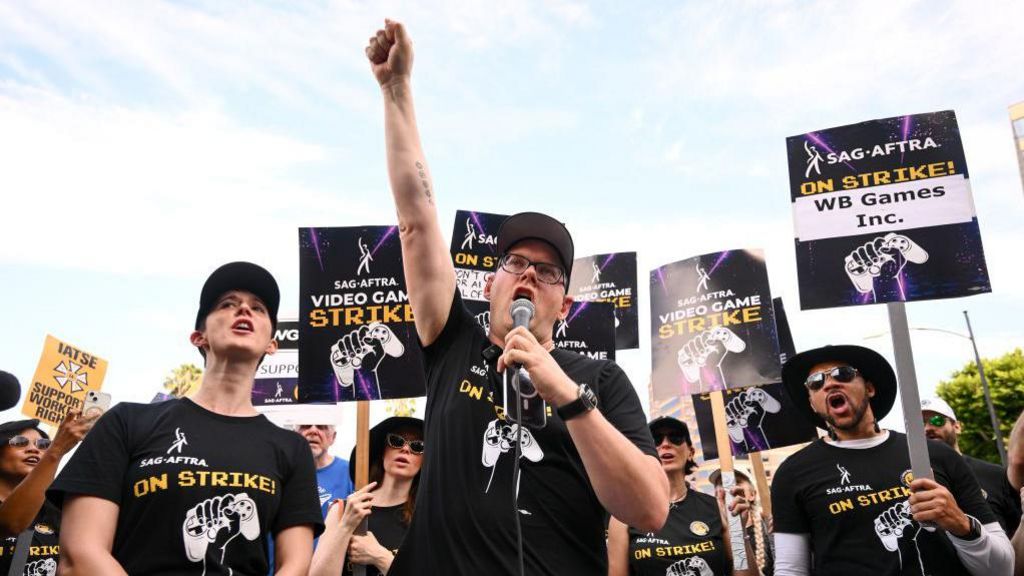 A man stands with his fist raised as he speaks into a microphone. He's wearing a black t-shirt with a picture of a fist clenched around a gaming controller and the text 