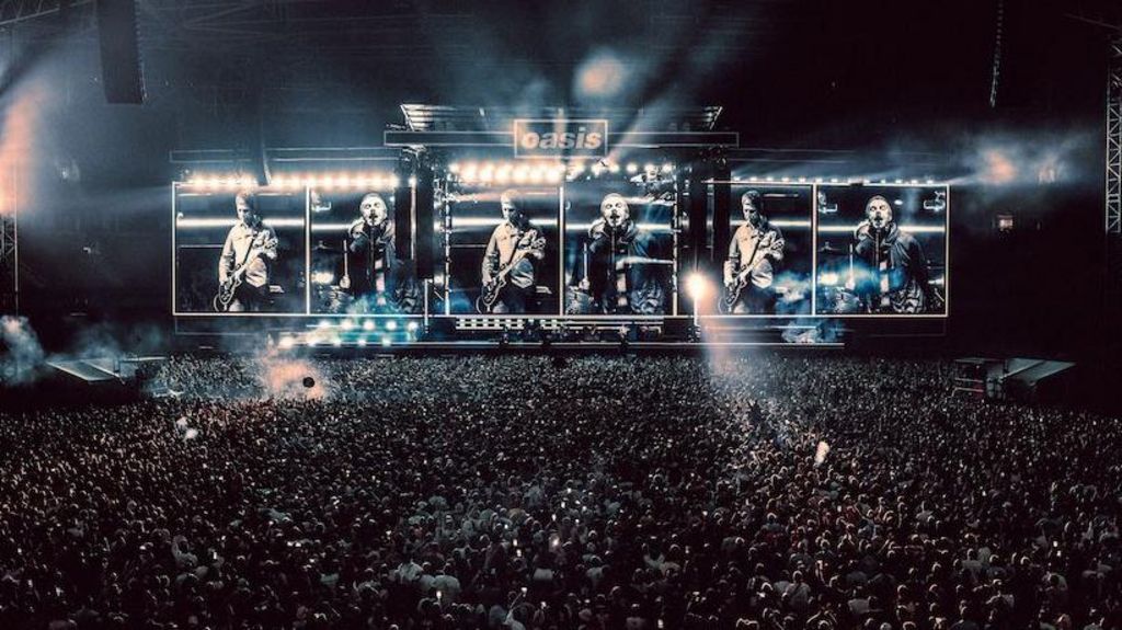 Wide shot of Oasis on stage in Cardiff. Thousands of people can be seen standing in front of the stage. Pictures of Liam and Noel performing can be seen on the stage screens.