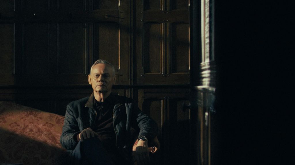 Clunes sat on a red sofa. He wears a brown jumper and black coat