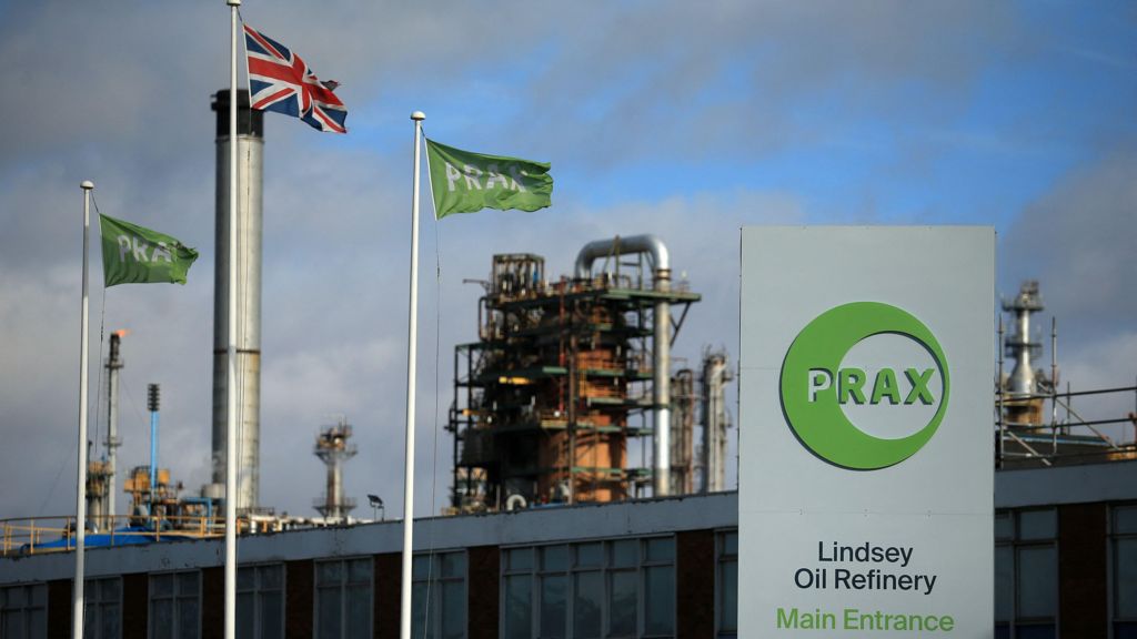 A metal structure in the background with a board on the left reading: 'PRAX. Lindsey Oil Refinery. Main Entrance'. There is a union jack flag on the left between two green flags with PRAX written.
