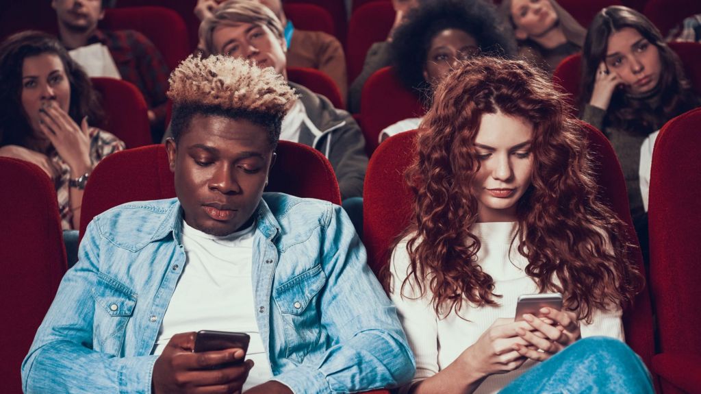 Young man and woman on their phones in the cinemam both in denim and white T shirts.