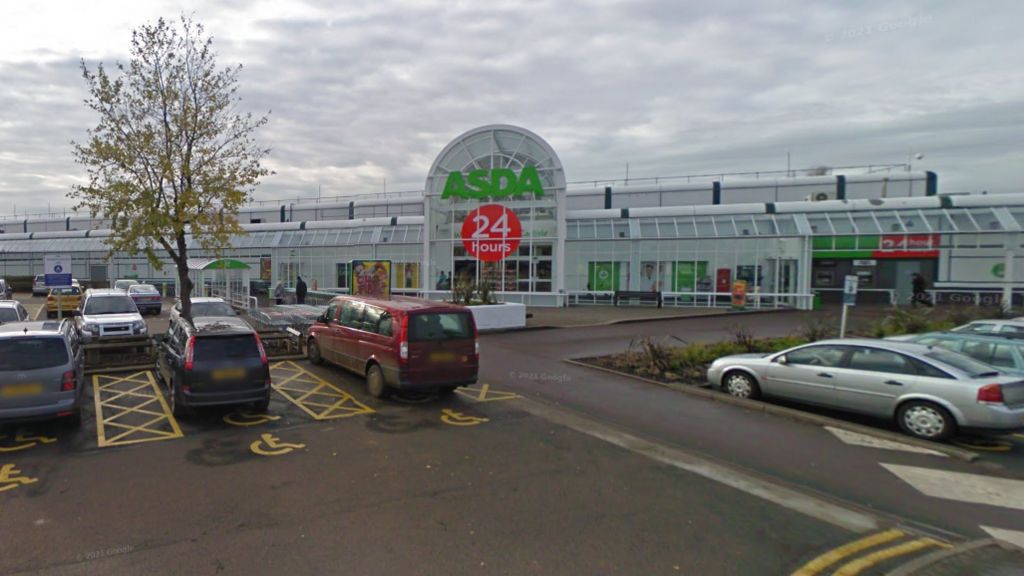 An Asda superstore with a glass, arched front which stretches the width of the frame. The green Asda sign can be seen alongside a red circle which reads '24 hours'. A busy car park sits in front of the store.