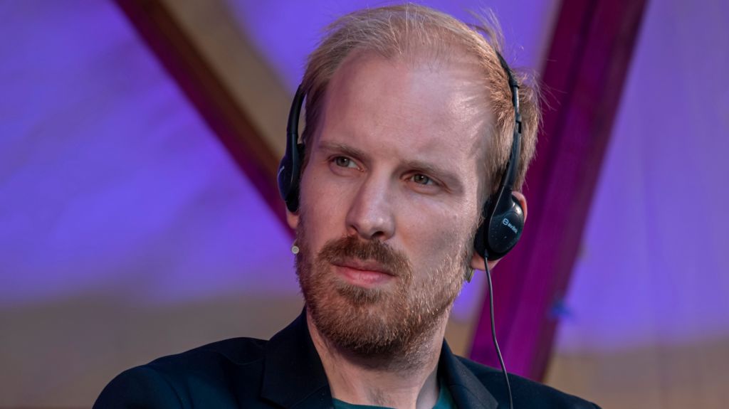 Rutger Bregman, wearing headphones, is looking to his right with a thoughtful expression - he is wearing a dark suit that can just be seen, he has blond-red hair and a short beard and moustache - he is inside a building with purple lighting behind him 