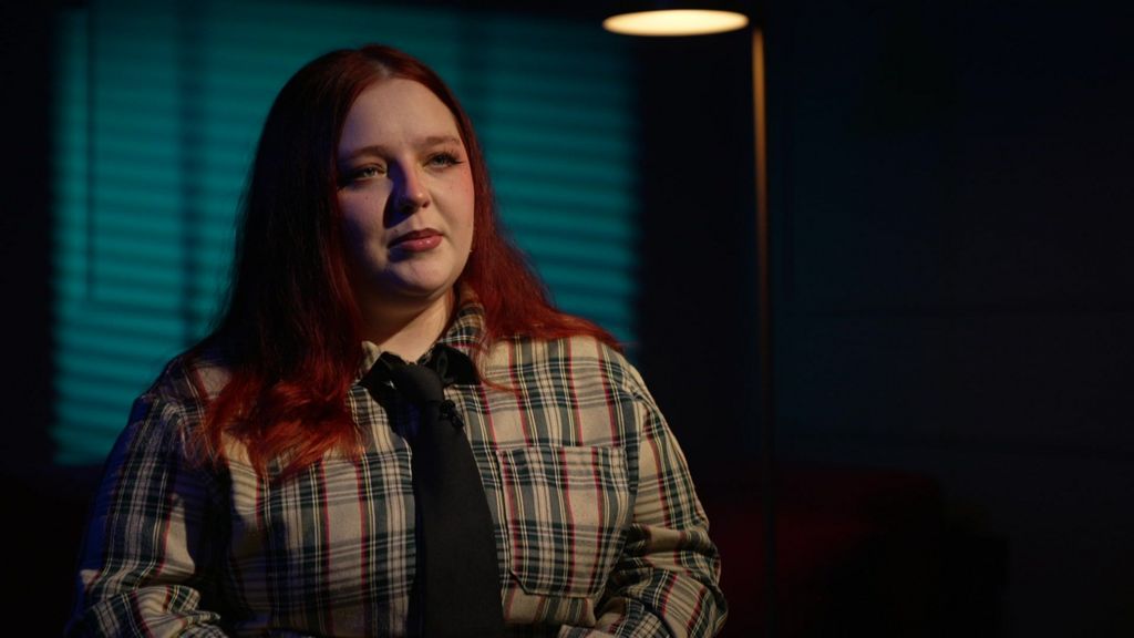 Amia Rohleder‑Cook sits during a filmed interview, wearing a checked shirt and tie, with blue lighting in the background.