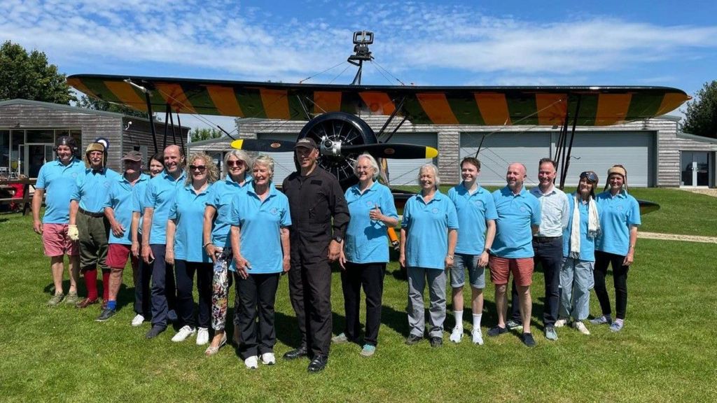 The "flying circus" stand in front of a biplane wearing light blue t-shirts and the pilot is dressed in black and is standing in the middle of the group. 