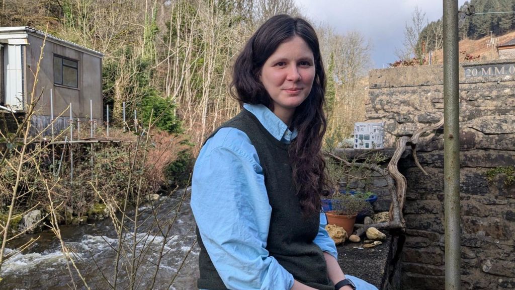 Georgie Scott, wearing a light blue shirt and knitted waistcoat, with long brown hair, sits on a garden wall by a river, looking into the camera. She is surrounded by trees.