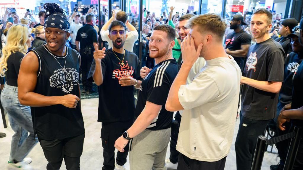 KSI, Vikram Singh Barn, Ethan Payne, Harry Lewis, Joshua Bradley, TBJZL and Simon Minter inside a shop - Lots of fans are seen in the background, filming on their phones from outside the store.