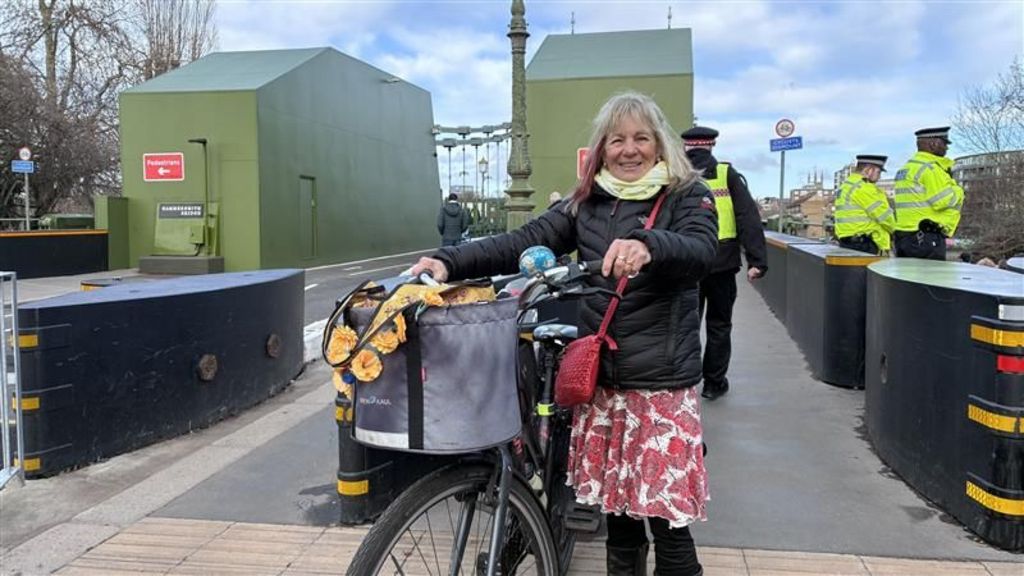 Ruth Mayorcas, wearing a black puffer jacket, red and white skirt, and is keeping her bike to one side, behind her is the walkway over Hammersmith bridge and some police officers and black bollards. 