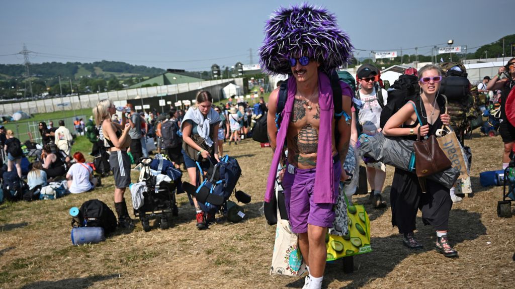 Man in a fluffy purple hat, scarf and shorts walking across festival site with fields in the distance