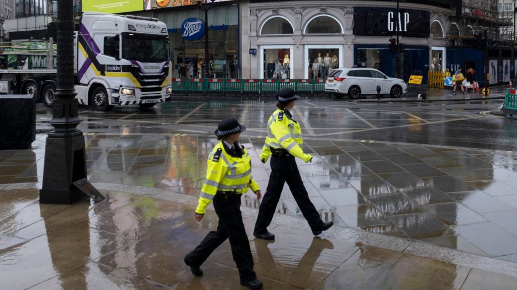Two uniformed police officers walk across a rain-soaked pavement in London, with shops, a white car, and a large lorry visible in the background.