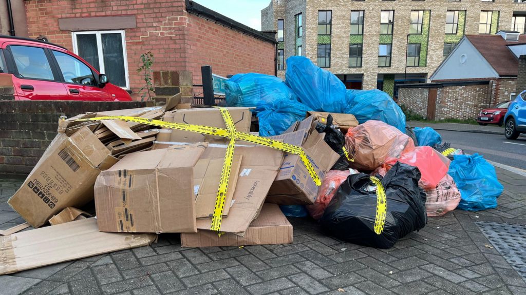 Cardboard boxes and blue, orange and black refuge bags are piled on a street corner. They have yellow environmental crime scene tape stuck across the mess