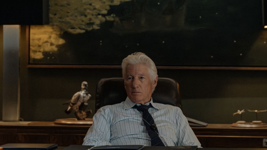 Richard Gere sits on an office chair wearing a tie and blue stripe shirt
