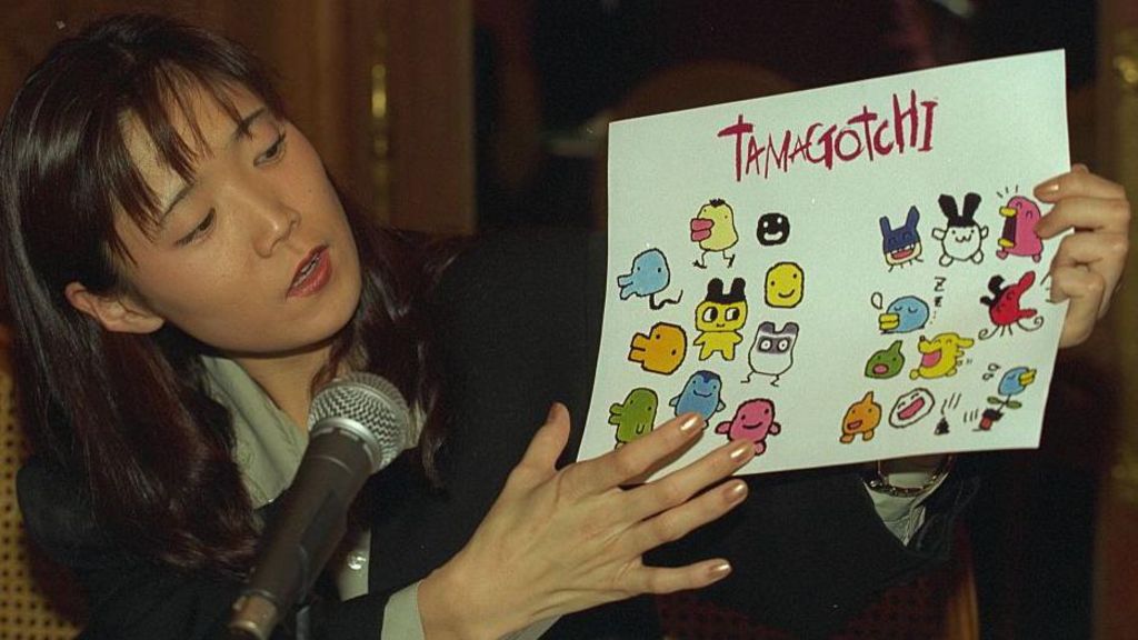 A Japanese woman stands on stage, holding a sign reading Tamagotchi and featuring many of the small, cute designs in the toy