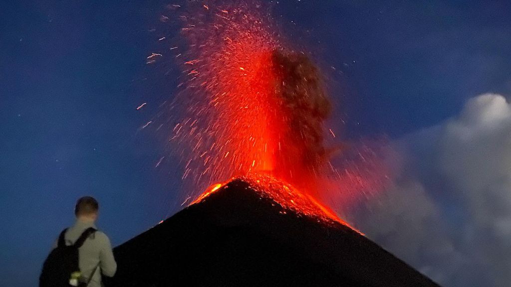 Tourists look not far from an erupting volcano, which is spewing red and orange lava