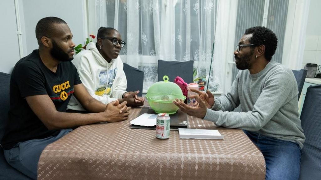 Three people sit at the kitchen table discussing. There is a brown table mat and a green container containing food.