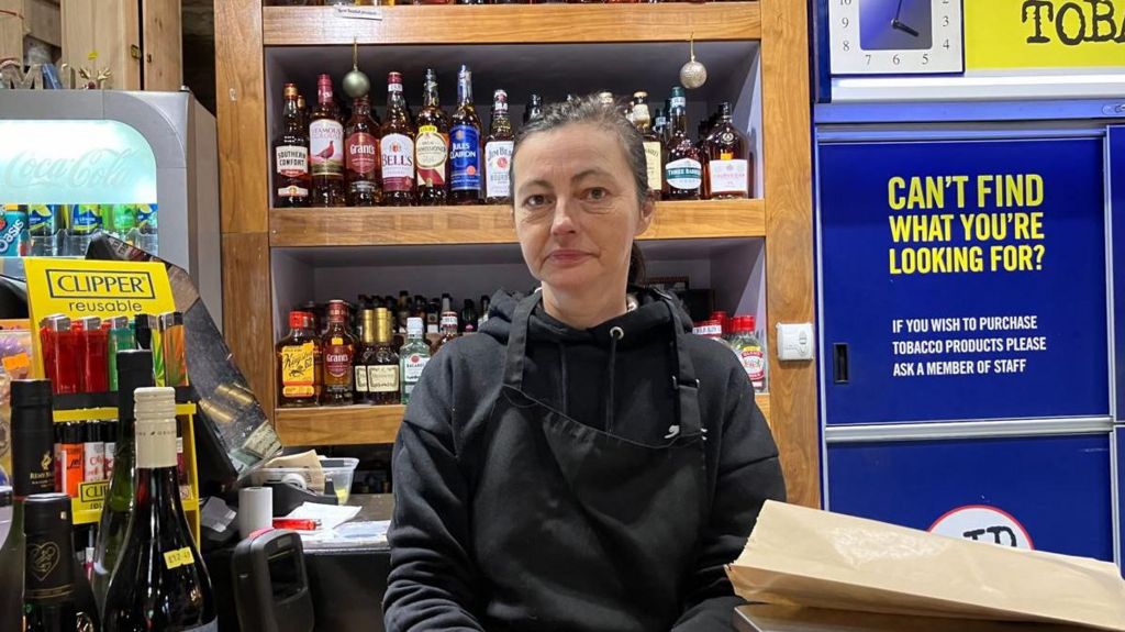 A middle-aged woman with dark hair in a ponytail wears a black apron and stands behind the counter in a shop with a host of alcohol on shelves behind her. 