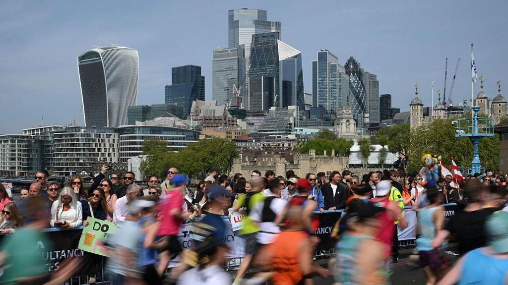 A blurred motion shot of runners passing, with the modern skyscrapers of the London city skyline in the background.