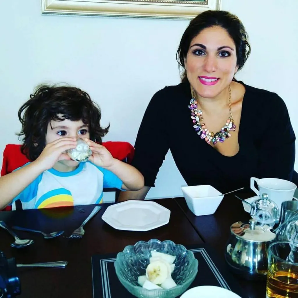 Vicki and one of her children sitting at a dining table set with plates, cutlery, a bowl of sliced fruit, a teapot, and a glass jug. The child is wearing a T‑shirt with a rainbow design and she is wearing a black top with a colourful beaded necklace. The table is black glass and the wall behind them is white and has a framed picture.