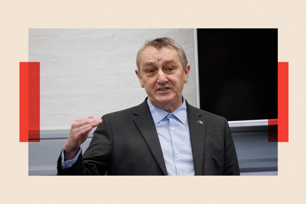Wales Green Party Leader, Anthony Slaughter. He is wearing a dark suit and a blue shirt buttoned up at the collar. He raises his right hand as if for emphasis. Behind him a white wall and a blackboard are visible.