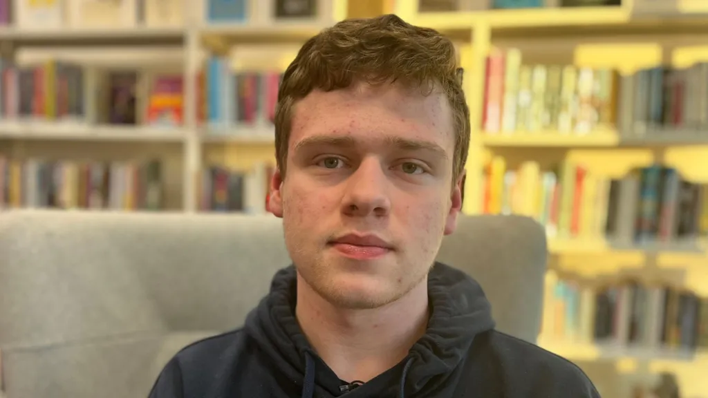 Liam has short, brown, wavy hair and is wearing a grey hoody. He is sitting in a  grey high-backed armchair in a library and looking directly into the camera.  