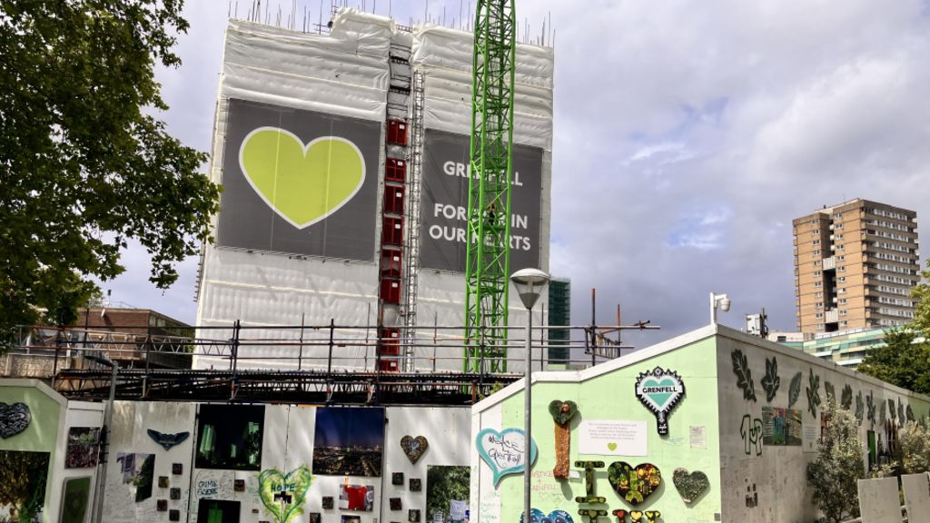 A partially demolished Grenfell tower with a wall surrounding the area which is covered in tributes and drawings. The building is covered in a wrap which features a green heart.