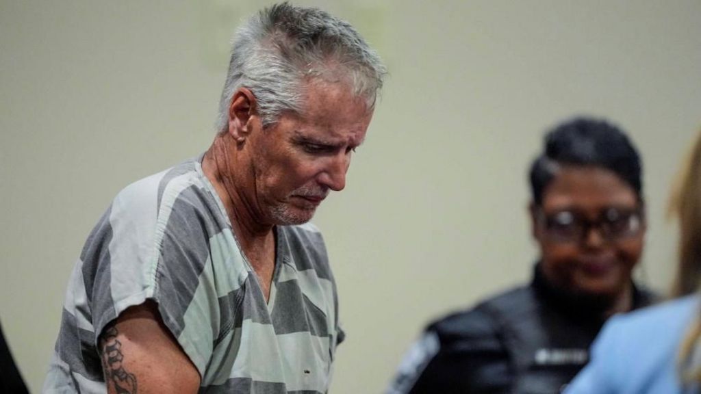 Colin Gray in court wears a dark and pale grey stripped prison uniform. He has grey close cropped hair and looks down.