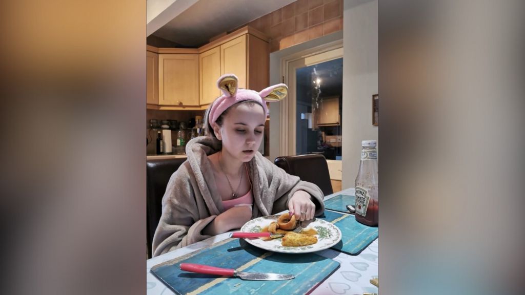 Lucy Dare as a young teenager, sat at a dining table in front of a small plate of food, which she is picking up in her left hand. She is wearing a dressing gown and a pink headscarf.