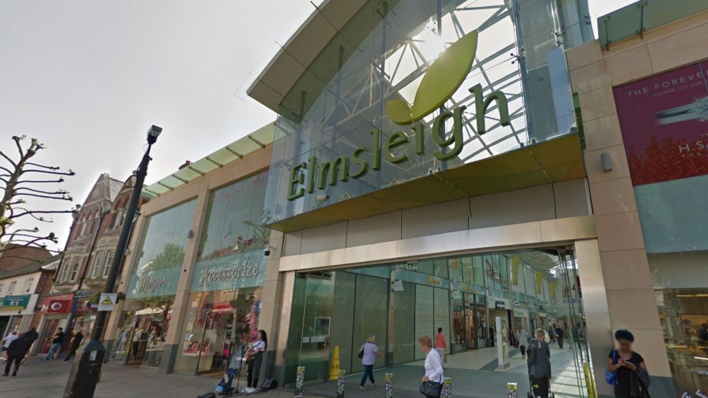 Outside of a shopping centre. There are people walking in and out of the entrance. Above the entrance is lots of glass, on which there is the word Elmsleigh written in green lettering