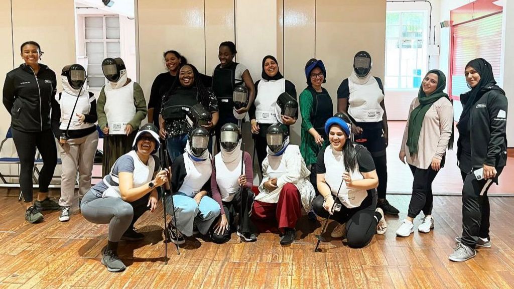 A group of women, some in fencing gear, stand in a room with wooden flooring