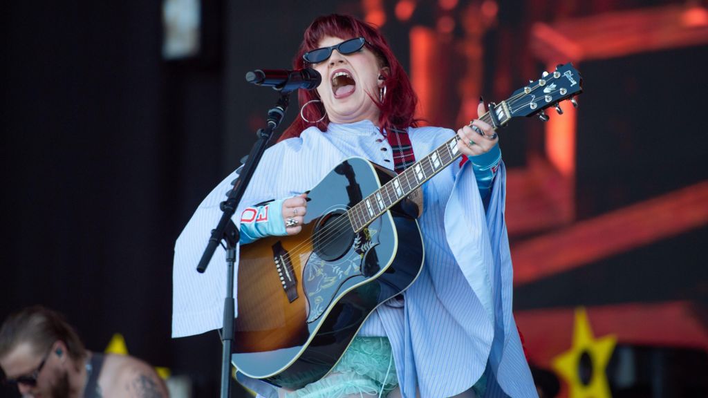 CMAT sings, wearing black sunglasses and a blue cape, her hair red, into a microphone while holding a guitar.