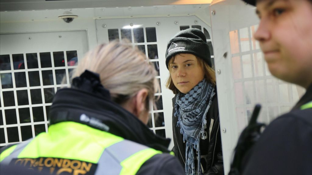 Greta Thunberg inside a police vehicle, looking through a partition, with police officers visible in the foreground.