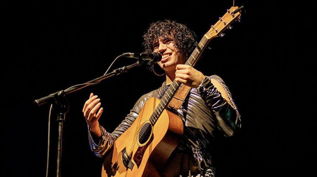 Jacob Alon is holding a guitar and singing into a microphone. They are wearing a patterned outfit. 