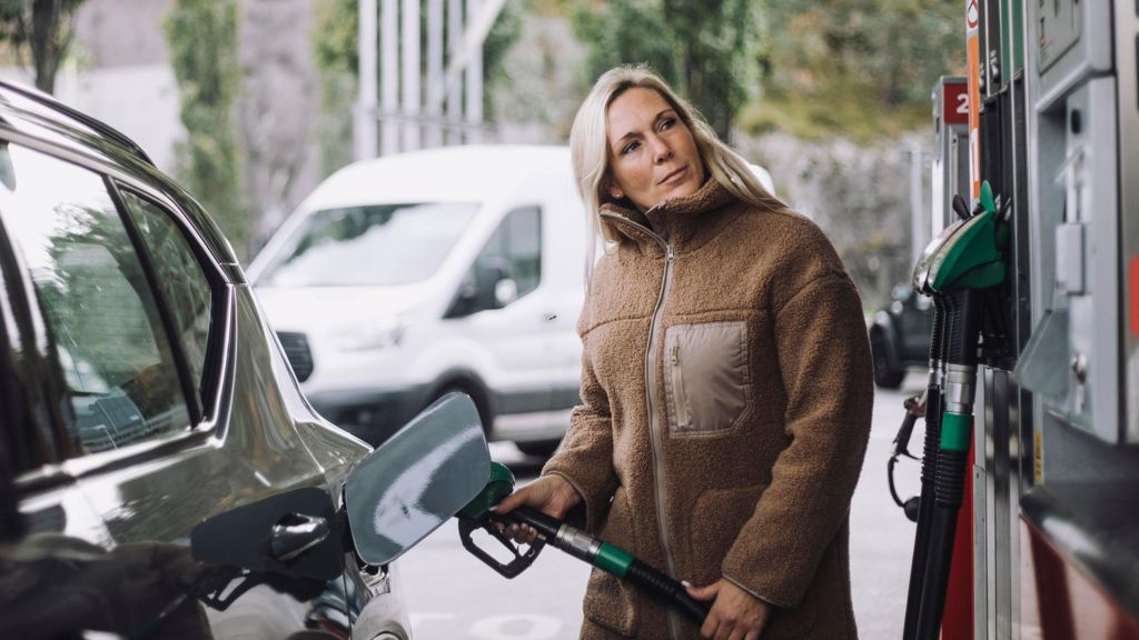 A stock photo of lady in a brown jacket filling up her car with fuel at a petrol station.
