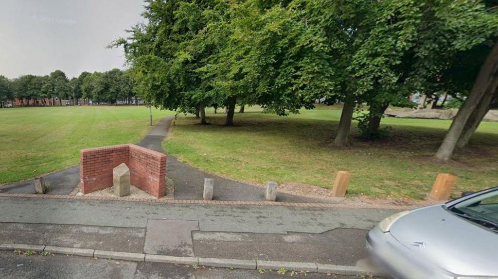 The edge of Peace Park, with a silver car parked on a street, close to bollards at the end of a pathway