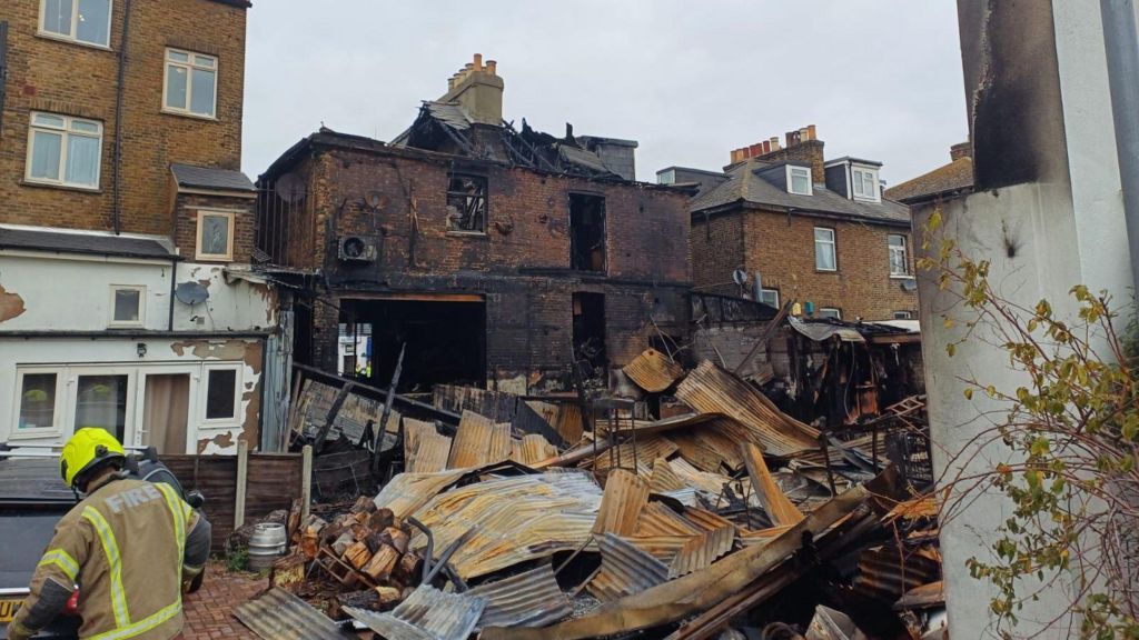 A firefighter stands in front of a burnt out brick building and a sea of rubble and debris