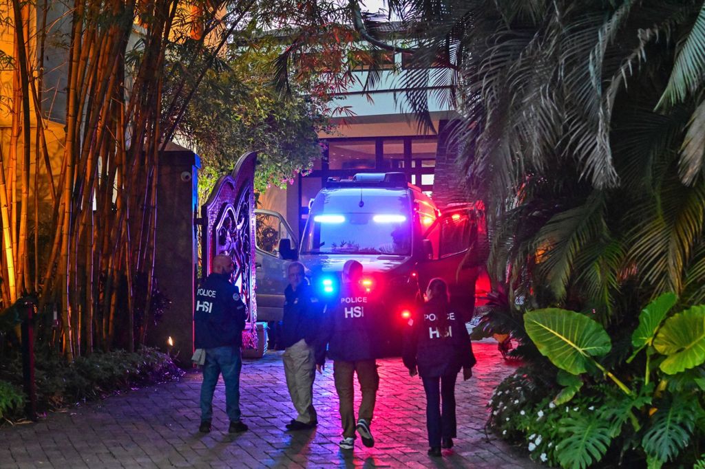 Police HSI officers walking toward a building at night, with a police vehicle flashing lights nearby, surrounded by greenery and illuminated by various lights.